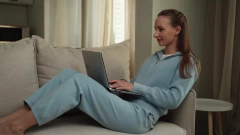 Young brunette uses a laptop while sitting on the couch in the living room at Stock Footage 262631113