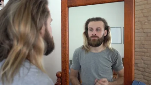 Young Brutal beard man in a T-shirt  is looking in a mirror twist out his hair Stock Footage 147895059