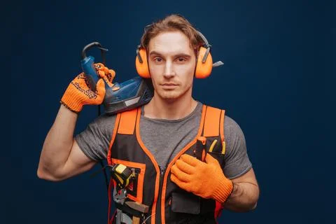 Young builder is serious while having equipment in arm, isolated over blue Stock Photos