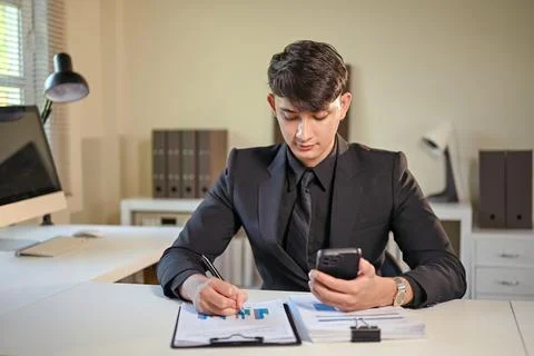 Young businessman analyzing financial reports while checking smartphone at 스톡 사진