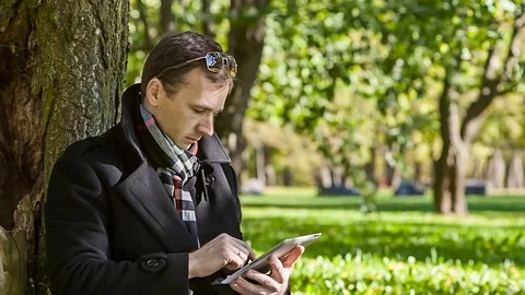 Young businessman in a coat with tablet computer sitting in the park Stock Footage 79106761