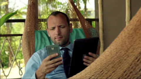 Young businessman comparing data on smartphone and tablet computer on hammock Stock Footage 53232822