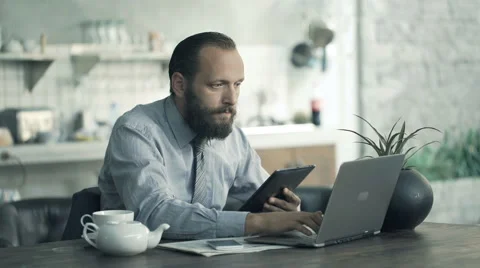Young businessman comparing data on tablet and laptop by table in kitchen Stock Footage 64056834