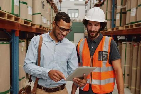 Young businessman showing data in touchpad to warehouse worker wearing hardhat Stock Photos