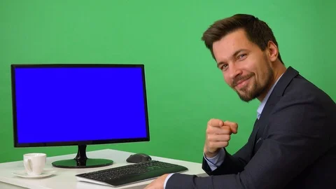 A young businessman sits in front of a computer with blue screen and points at Stock Footage 115124339