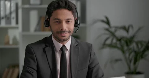 Young businessman sitting at the office, using headphones, looking at camera. Video stock 248966324