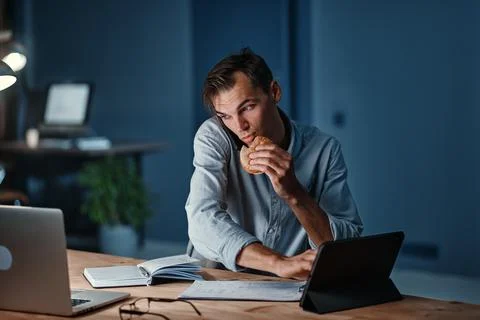 Young businessman snacking on a hamburger while working on a new project. Stock Photos