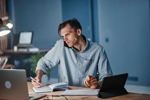Young businessman snacking on a hamburger while working on a new project. Stock Photos