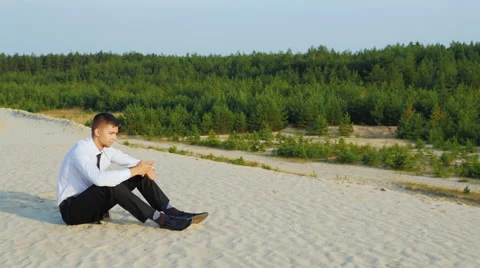 Young businessman typing a message on your phone, sitting in the sand against Stock Footage 65987847