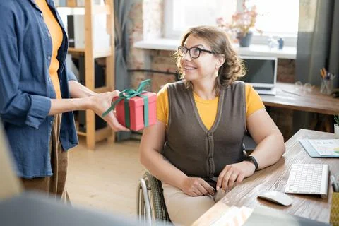 Young businesswoman in wheelchair looking at colleague passing her packed gift Stock Photos