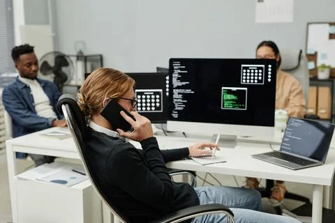 Young busy programmer talking on mobile phone while sitting in front of computer 스톡 사진