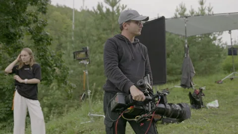 Young cameraman stands with camera in hand enjoying brief pause between actios Видео 303055398