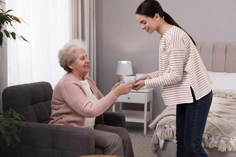 Young caregiver giving tea to senior woman in room. Home care service Stock Photos