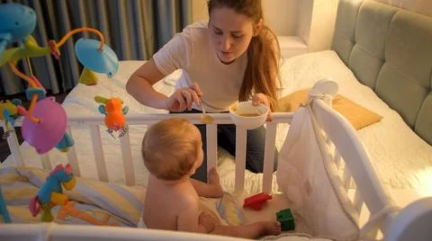 Young caring mother feeding her baby son with soup in baby crib at night Stock Photos