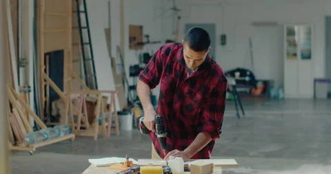 Young carpenter working in his workshop and drilling a hole on the plank with an Stock Footage 199617849