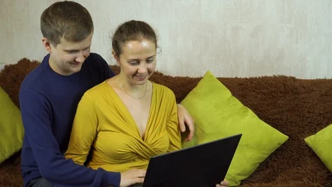 Young casual couple using laptop sitting on couch at home, 스톡 동영상 99402447