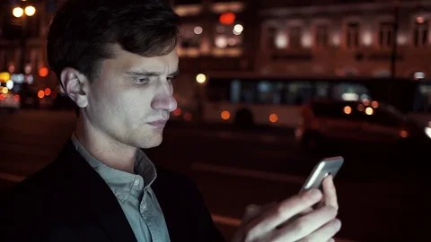 Young casual dressed man browsing his cellphone on a busy urban street at night Stock Footage 80009727