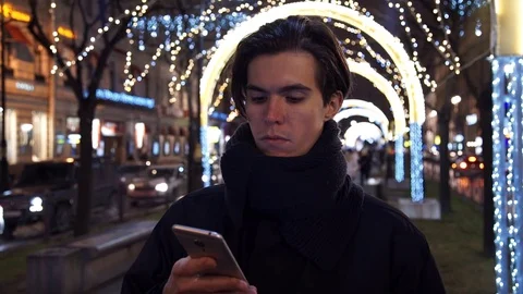 Young casual dressed man browsing his cellphone on a busy urban street at night Stock Footage 83630035