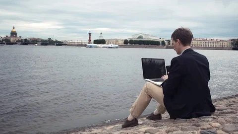 Young casual dressed man using laptop sitting on embankment. Working outdoors Stock Footage 80009061