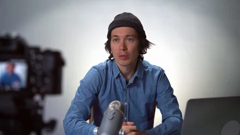 Young casual man blogger in hat and glasses looking at camera speaking recording Stock Footage 128711253