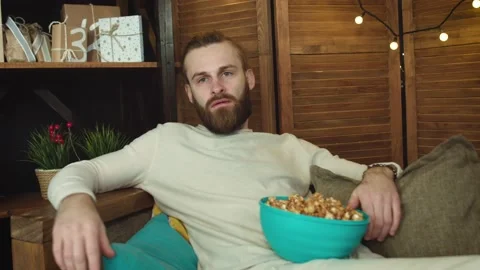 Young  Casual Man is watching something interesting on  TV eating popcorn Stock Footage 147034295