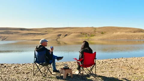 Young caucasian Couple sit on foldable chairs by lake drink coffee outdoors i Vídeos de archivo 165219070