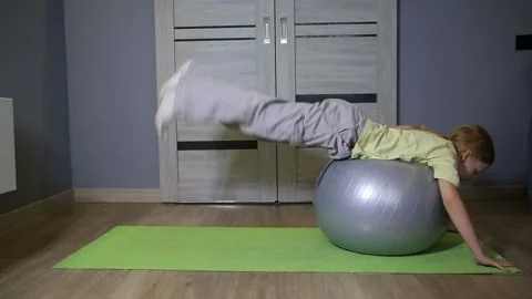 Young caucasian girl performing back exercises with fitness ball on green mat Stock Footage 327188597