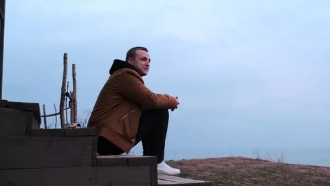 Young caucasian guy in beige outerwear, black jeans and white sneakers sits on w Stock Footage 126922810