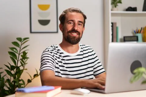 Young caucasian guy looking at camera smiling, sitting at home using laptop to Stock Photos