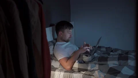 Young caucasian guy waking up at night sitting on bed looking at computer screen Stock Footage 145494500