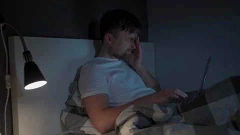Young caucasian guy waking up at night sitting on bed looking at computer screen Stock Footage 145494827