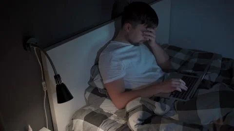 Young caucasian guy waking up at night sitting on bed looking at computer screen Stock Footage 145495027