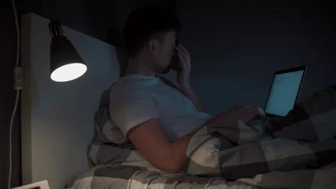 Young caucasian guy waking up at night sitting on bed looking at computer screen Stock Footage 145495229