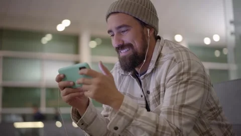 Young Caucasian laughing man having fun watching video with earphones on cell Stock Footage 256092582