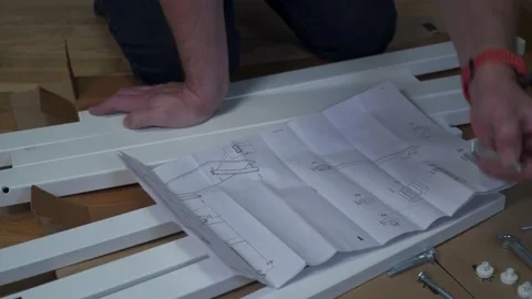Young Caucasian man assembles white furniture from flat box using instructions Stock Footage 150770126
