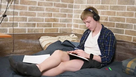 Young Caucasian Man Sitting on Bed with Headphones, Drawing in Sketchbook Stock Footage 340458144
