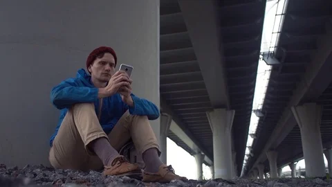 Young caucasian man using smartphone sitting on ground under road bridge Stock Footage 80285894