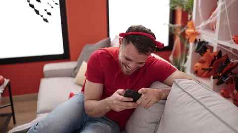 Young caucasian man wearing devil costume sitting on the sofa using smartph.. Stock Footage 234155676