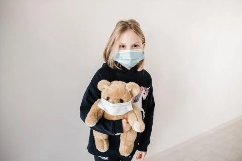 Young caucasion girl wearing a mask and holding a teddy bear who is also wear Stock Photos