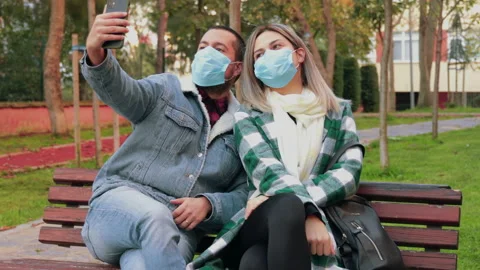 Young charming couple sitting on a bench in park, wearing protective masks Stock Footage 143715136