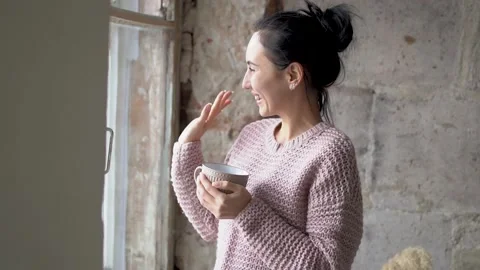 Young charming girl stands at the window and drinks coffee, smiles and dreams Vi Stock Footage 169335136