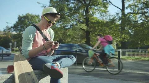 Young cheerful boy sitting on a bench and texting on a mobile phone, outdoors. 動画素材 42990409