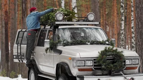 Young cheerful man in blue jacket holding mini Christmas tree near trunk old car Stock Footage 259303291