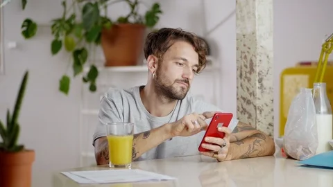 Young cheerful man using red smartphone device drinking fresh orange juice at Stock Footage 108797993