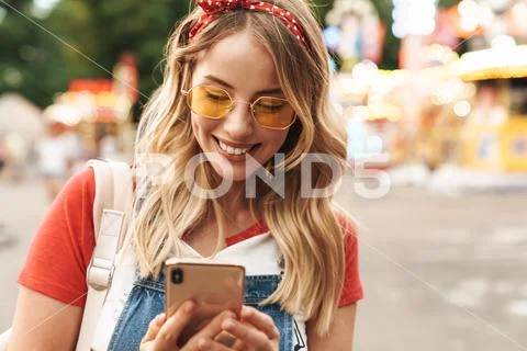 Young cheery blonde woman in amusement park using mobile phone. Stock Photos