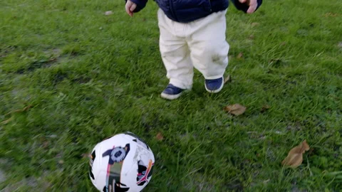 Young child in blue jacket and white pants engages with a soccer ball on grassy Video stock 327851236