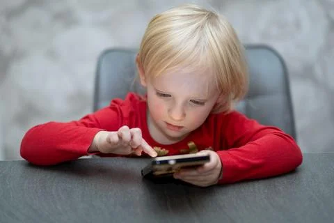 Young child focused on smartphone while seated at a table in a bright indoor Stock Photos