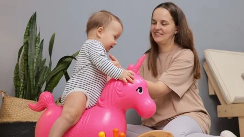 A young child joyfully rides a bright pink unicorn toy while a smiling mother Vídeo Stock 299939640