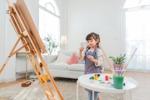Young child paints on table while standing near easel in bright living room.. Stock Photos