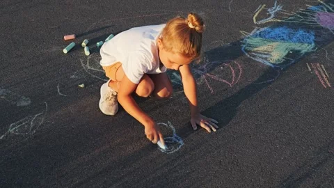 A young child sits on the ground and draws with chalk on the pavement. It is a Vídeo Stock 340960507